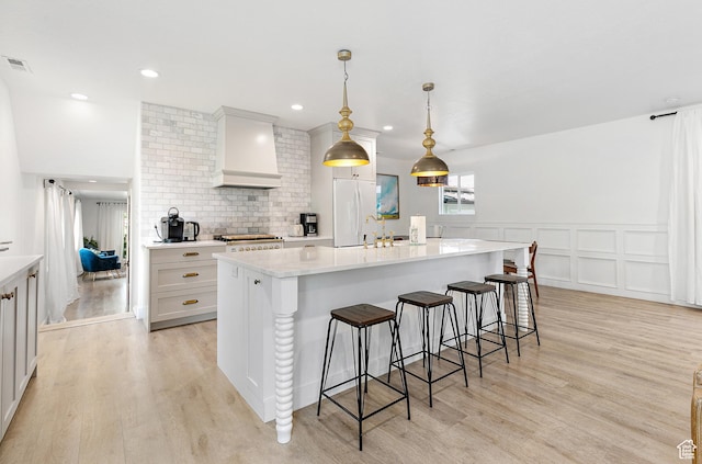 kitchen with wall chimney exhaust hood, light wood-style floors, a kitchen bar, a kitchen island with sink, and wainscoting