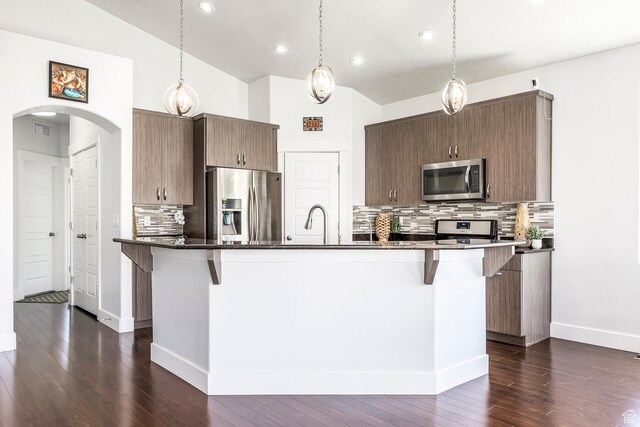 kitchen featuring appliances with stainless steel finishes, arched walkways, a kitchen bar, decorative backsplash, and dark wood-style floors