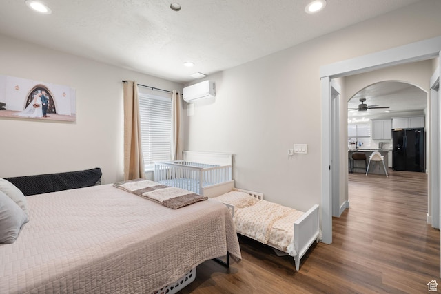 bedroom with black fridge with ice dispenser, a wall mounted AC, wood finished floors, recessed lighting, and arched walkways