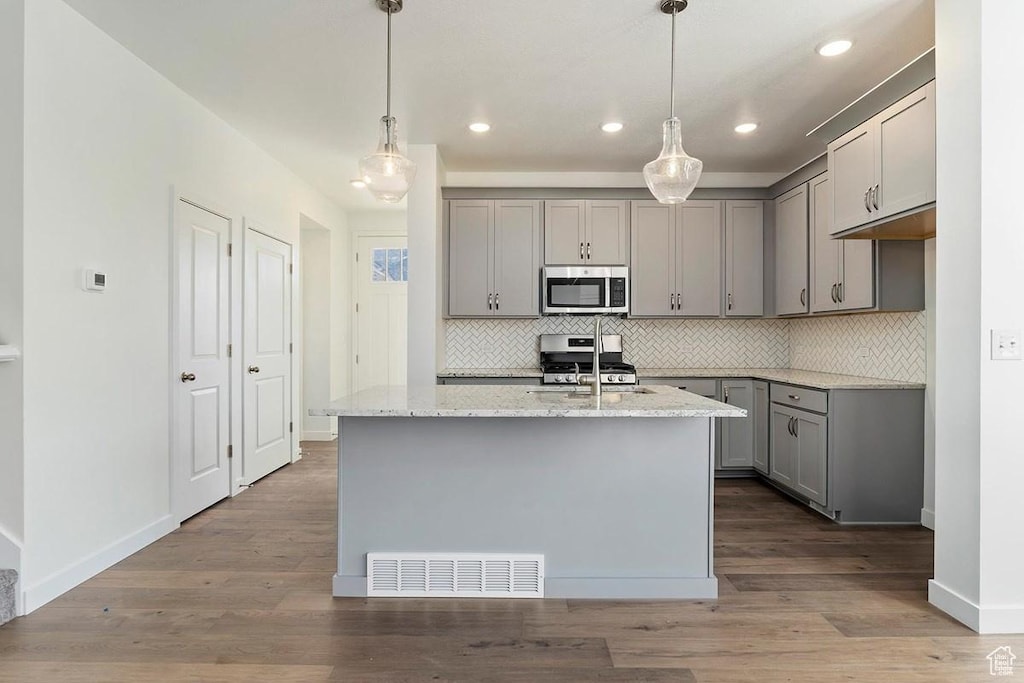 kitchen featuring appliances with stainless steel finishes, gray cabinetry, backsplash, light stone counters, and a kitchen island with sink