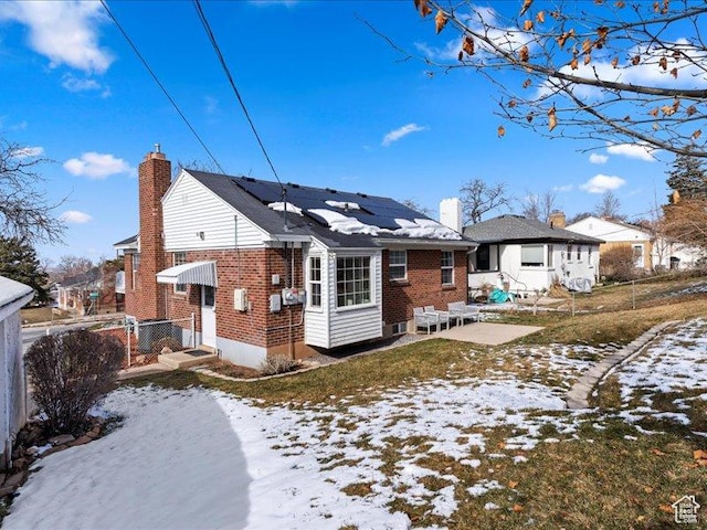 snow covered back of property with solar panels, brick siding, a chimney, fence, and a patio