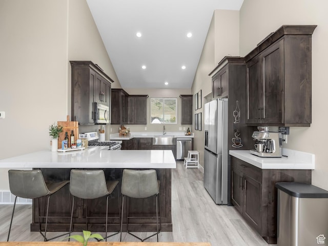 kitchen featuring appliances with stainless steel finishes, a peninsula, dark brown cabinets, light wood-style floors, and a kitchen bar