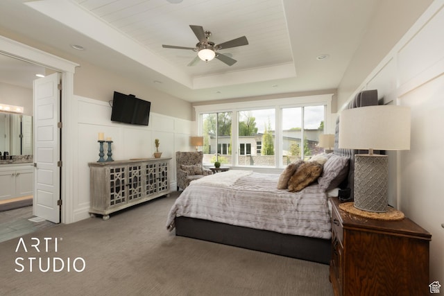 bedroom with a tray ceiling, carpet, a decorative wall, ensuite bath, and a ceiling fan