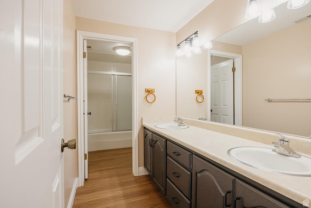bathroom featuring wood finished floors, double vanity, bath / shower combo with glass door, and baseboards