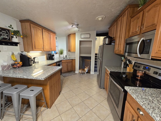kitchen featuring appliances with stainless steel finishes, brown cabinets, light stone counters, a peninsula, and a textured ceiling
