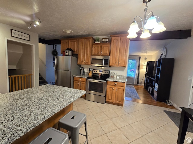 kitchen with appliances with stainless steel finishes, light tile patterned floors, a chandelier, brown cabinetry, and light stone countertops