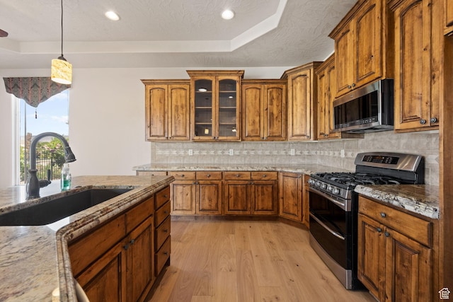kitchen with stainless steel appliances, a tray ceiling, a sink, light wood finished floors, and brown cabinets