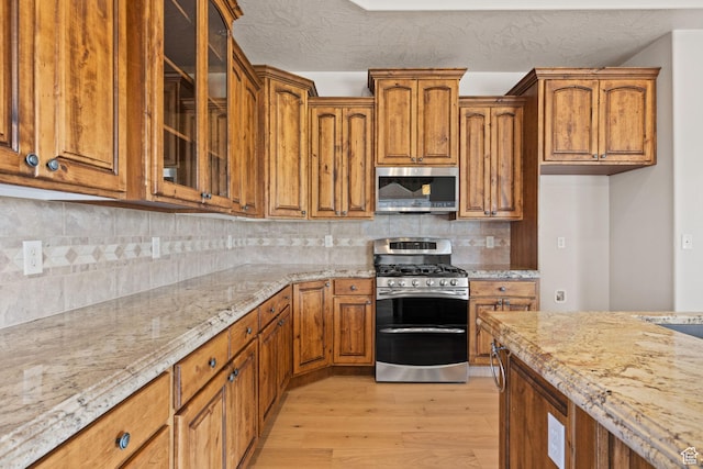 kitchen with appliances with stainless steel finishes, light wood-style flooring, light stone counters, brown cabinets, and glass insert cabinets