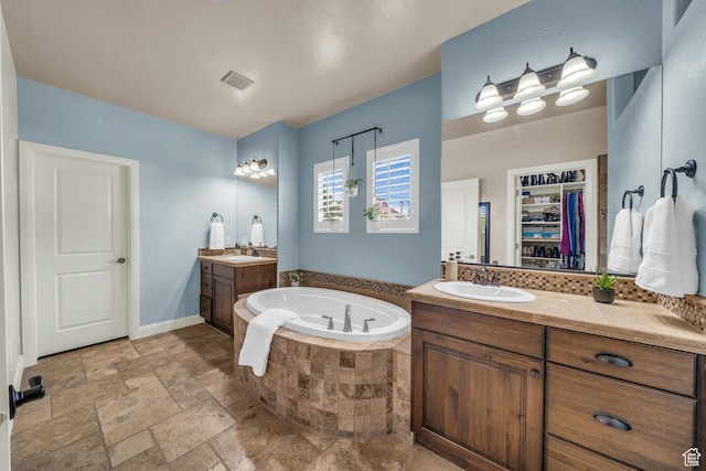 bathroom featuring a bath, two vanities, stone tile flooring, a walk in closet, and baseboards