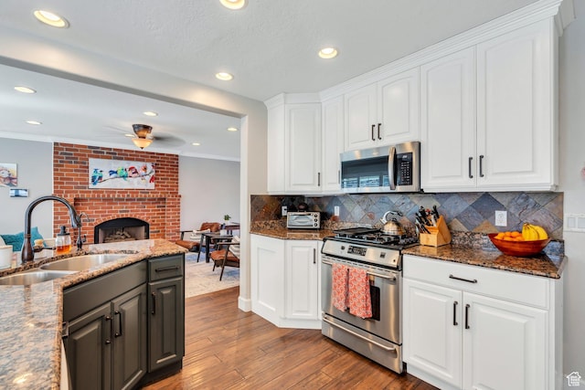 kitchen featuring stainless steel appliances, a sink, a brick fireplace, white cabinetry, and wood finished floors