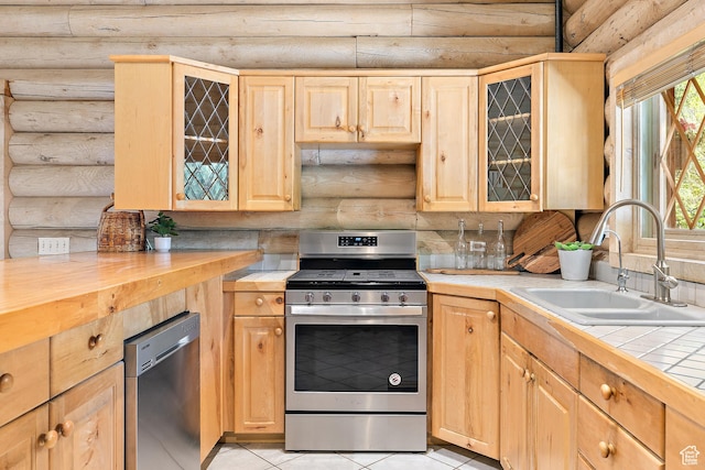 kitchen with stainless steel appliances, rustic walls, a sink, light brown cabinetry, and tile countertops