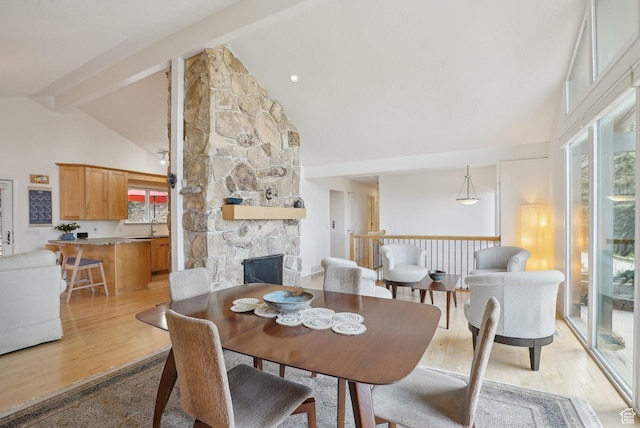 dining room with healthy amount of natural light, high vaulted ceiling, beam ceiling, a stone fireplace, and light wood-type flooring