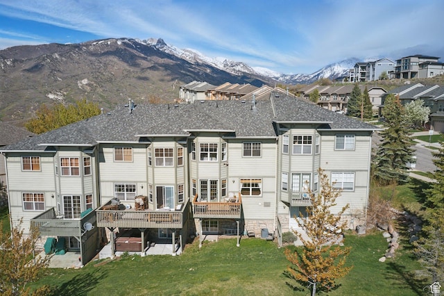 back of property with a lawn, a deck with mountain view, a shingled roof, and a patio