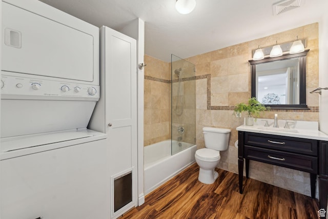 full bath featuring stacked washing machine and dryer, wood finished floors, vanity, toilet, and tile walls