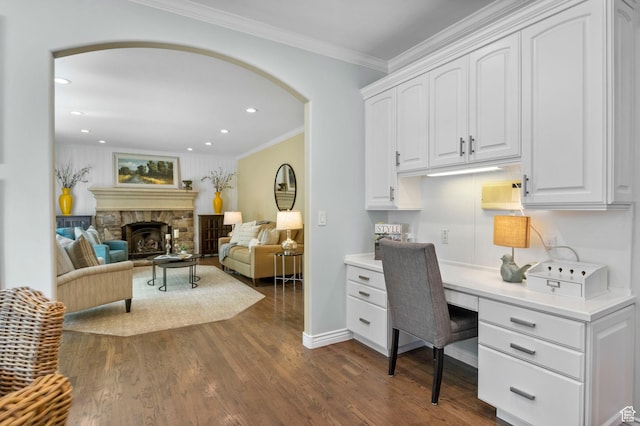 home office featuring arched walkways, built in desk, dark wood-style floors, ornamental molding, and a stone fireplace