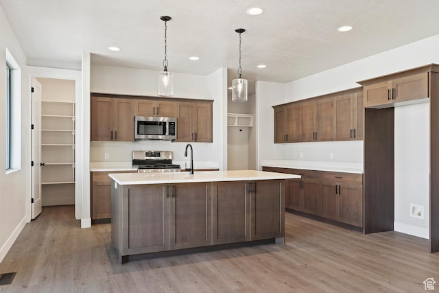 kitchen with stainless steel appliances, light countertops, light wood finished floors, an island with sink, and decorative light fixtures