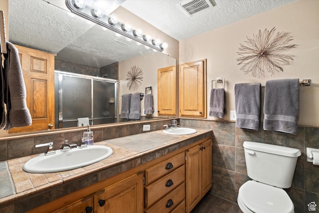 bathroom with a textured ceiling, a shower stall, toilet, double vanity, and tile walls