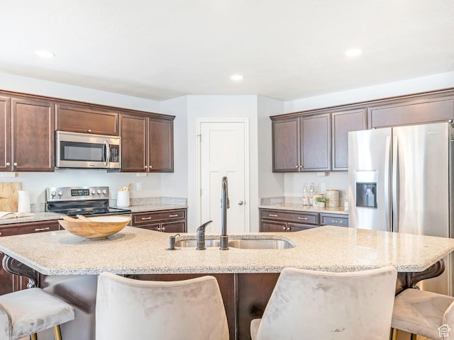 kitchen featuring appliances with stainless steel finishes, a sink, a breakfast bar, dark brown cabinets, and light stone counters