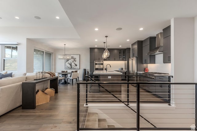 kitchen with wall chimney range hood, wood finished floors, recessed lighting, a center island with sink, and appliances with stainless steel finishes
