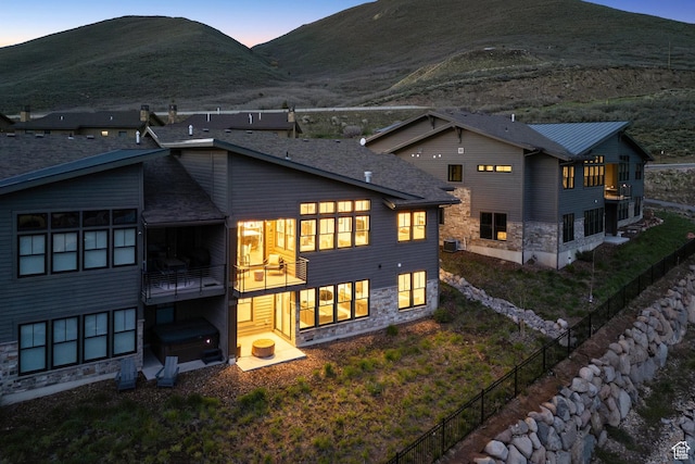 back of house at dusk with a balcony, a mountain view, stone siding, and fence