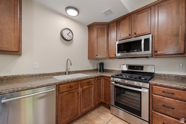 kitchen with appliances with stainless steel finishes, a sink, dark countertops, light tile patterned floors, and brown cabinetry