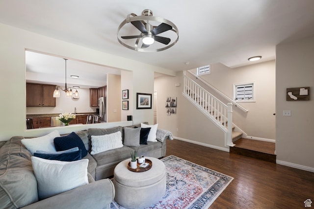 living room featuring ceiling fan, stairs, baseboards, dark wood-style floors, and a chandelier