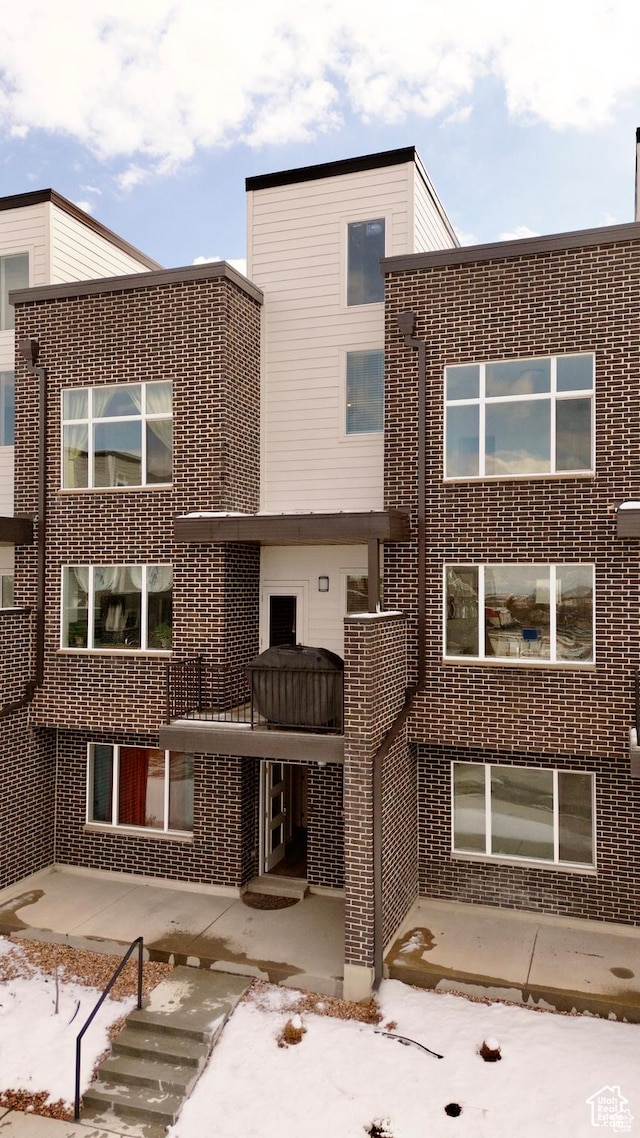 snow covered house with brick siding, a patio, and a balcony