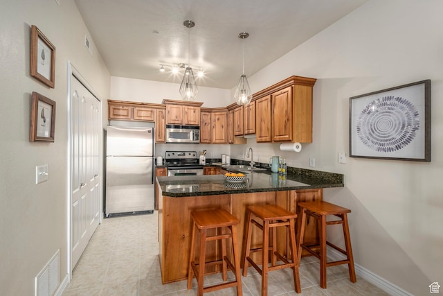 kitchen with appliances with stainless steel finishes, a peninsula, a sink, brown cabinets, and dark stone countertops