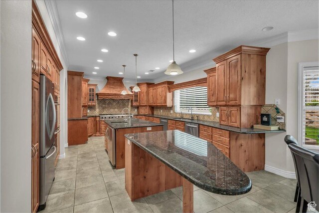 1659 S Cobblestone Ln, St George, UT 84790 - kitchen with an island with sink, custom range hood, brown cabinetry, appliances with stainless steel finishes, and decorative backsplash
