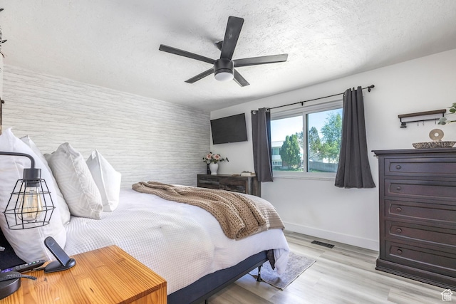 bedroom with baseboards, light wood-type flooring, a textured ceiling, a ceiling fan, and an accent wall