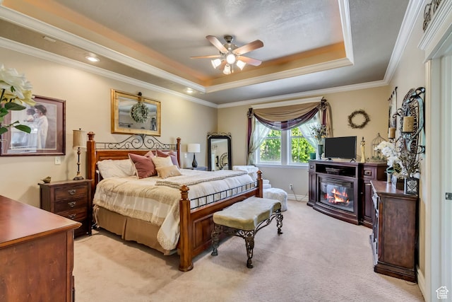 bedroom featuring a raised ceiling, light carpet, a glass covered fireplace, ornamental molding, and a ceiling fan