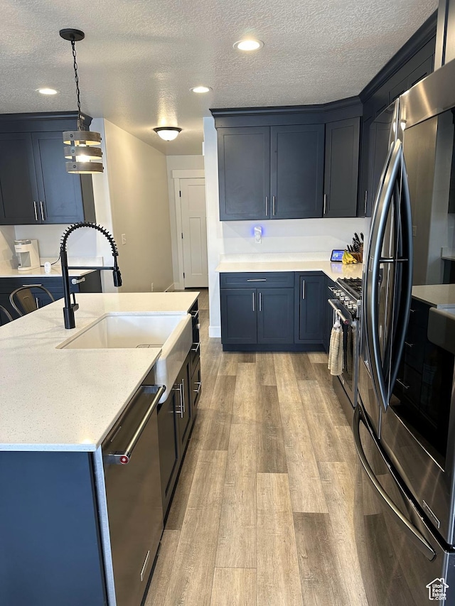 kitchen with stainless steel appliances, a sink, light wood finished floors, a textured ceiling, and hanging light fixtures