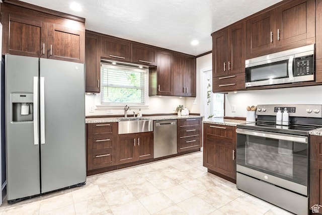 kitchen with stainless steel appliances, a sink, dark brown cabinets, light stone counters, and recessed lighting
