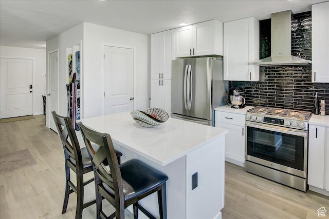 kitchen with appliances with stainless steel finishes, wall chimney exhaust hood, light wood-style floors, tasteful backsplash, and a kitchen breakfast bar