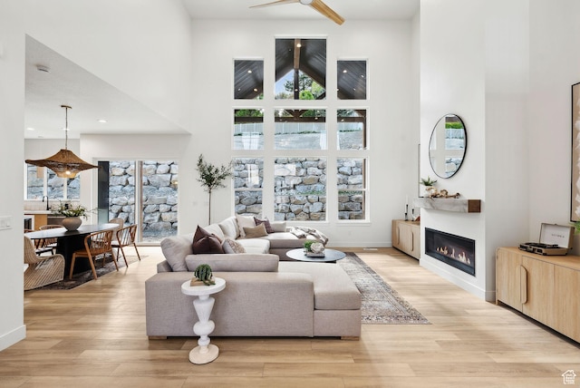 living area with a towering ceiling, a glass covered fireplace, healthy amount of natural light, light wood finished floors, and a ceiling fan