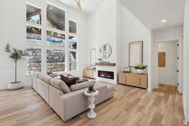 living room with high vaulted ceiling, light wood finished floors, a glass covered fireplace, baseboards, and recessed lighting