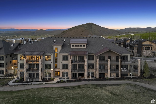 back of property at dusk featuring a mountain view, stone siding, and a shingled roof