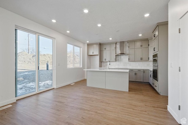 kitchen with wall chimney exhaust hood, gray cabinetry, stainless steel double oven, an island with sink, and tasteful backsplash