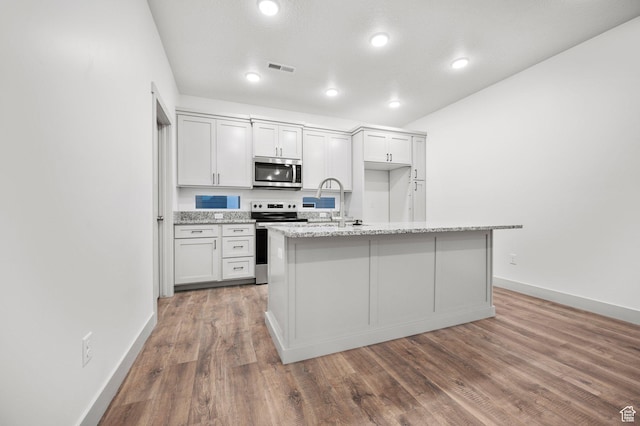 kitchen with stainless steel appliances, wood finished floors, a center island with sink, baseboards, and light stone counters