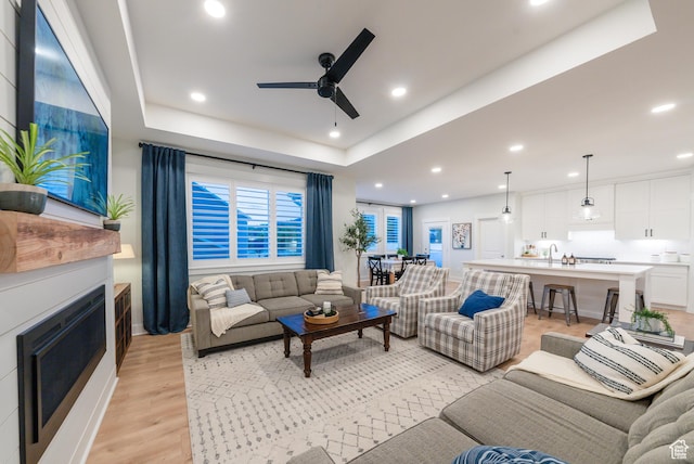 living area featuring ceiling fan, a raised ceiling, light wood finished floors, a fireplace, and recessed lighting