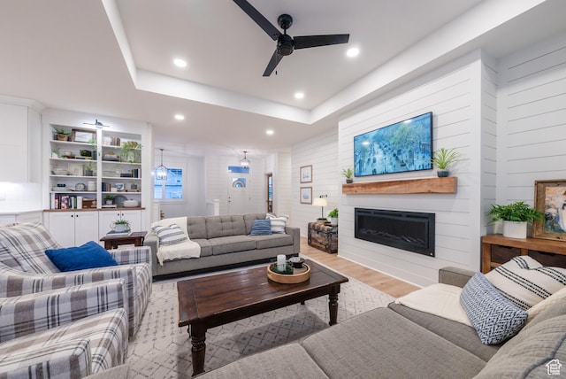 living area featuring a ceiling fan, light wood-style floors, a fireplace, a tray ceiling, and recessed lighting