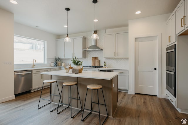 kitchen with stainless steel appliances, wall chimney range hood, a kitchen bar, wood finished floors, and recessed lighting