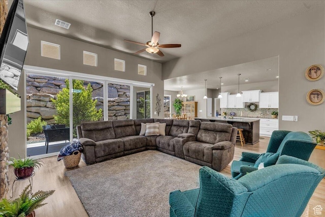 living area featuring a ceiling fan, a high ceiling, light wood-style floors, a textured ceiling, and a chandelier