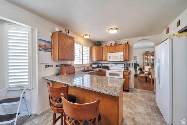 kitchen featuring white appliances, a peninsula, arched walkways, brown cabinets, and a sink