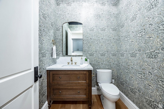 bathroom featuring wood finished floors, wallpapered walls, toilet, vanity, and baseboards