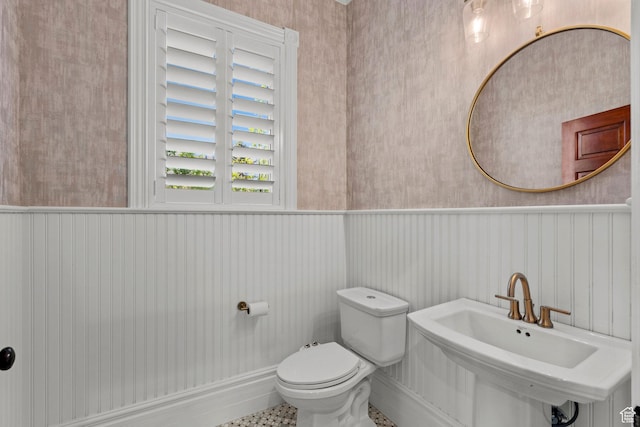 half bathroom featuring a wainscoted wall, toilet, a sink, and wallpapered walls