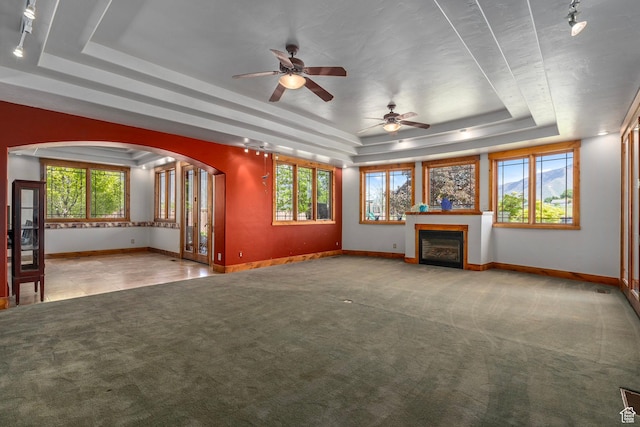 unfurnished living room featuring a tray ceiling, arched walkways, carpet floors, baseboards, and a ceiling fan
