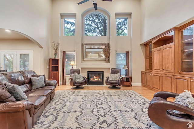 living room with a high ceiling, a ceiling fan, arched walkways, a glass covered fireplace, and light wood finished floors