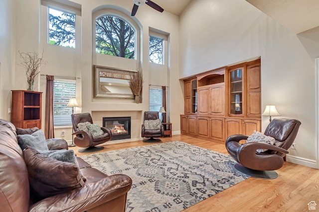 living area with a towering ceiling, light wood-style floors, a ceiling fan, a glass covered fireplace, and baseboards