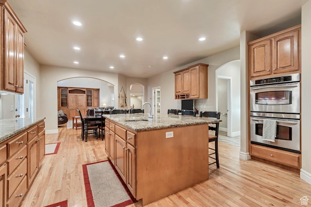 kitchen with arched walkways, stainless steel double oven, a sink, a kitchen island with sink, and a breakfast bar
